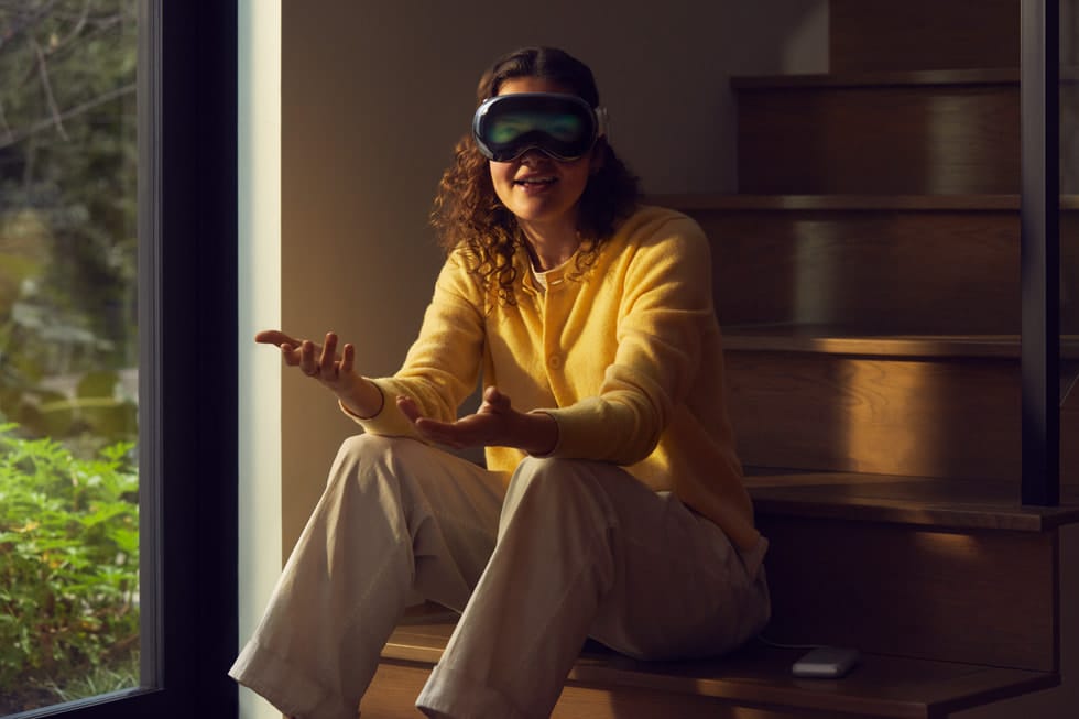 A woman wearing an Apple Vision Pro headset sits on the stares. The outside of the visor shows a dim, fuzzy image of her face that doesn't quite look right.