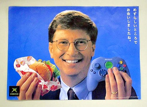 Xbox Japanese ad with Bill Gates smiling, holding up a hamburger in his right hand and an Xbox Controller S in his left hand.