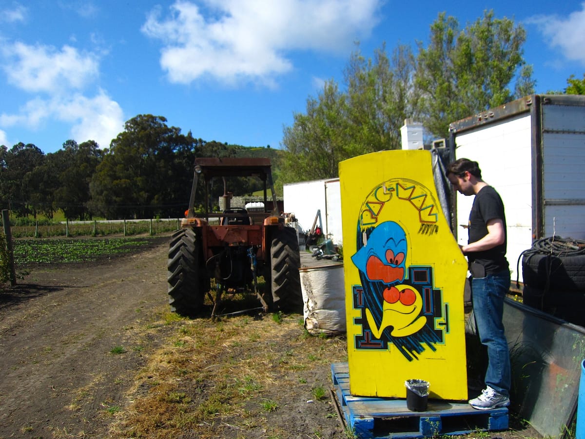 Photo of a farm with a tractor along a dirt path. There is a Pac-Man arcade machine with a man at the controls.