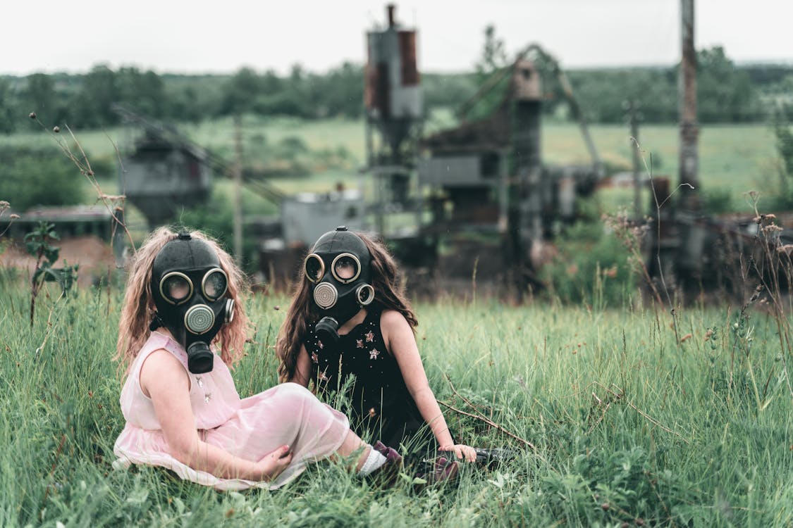 Two girls sit in a grassy field wearing gas masks.